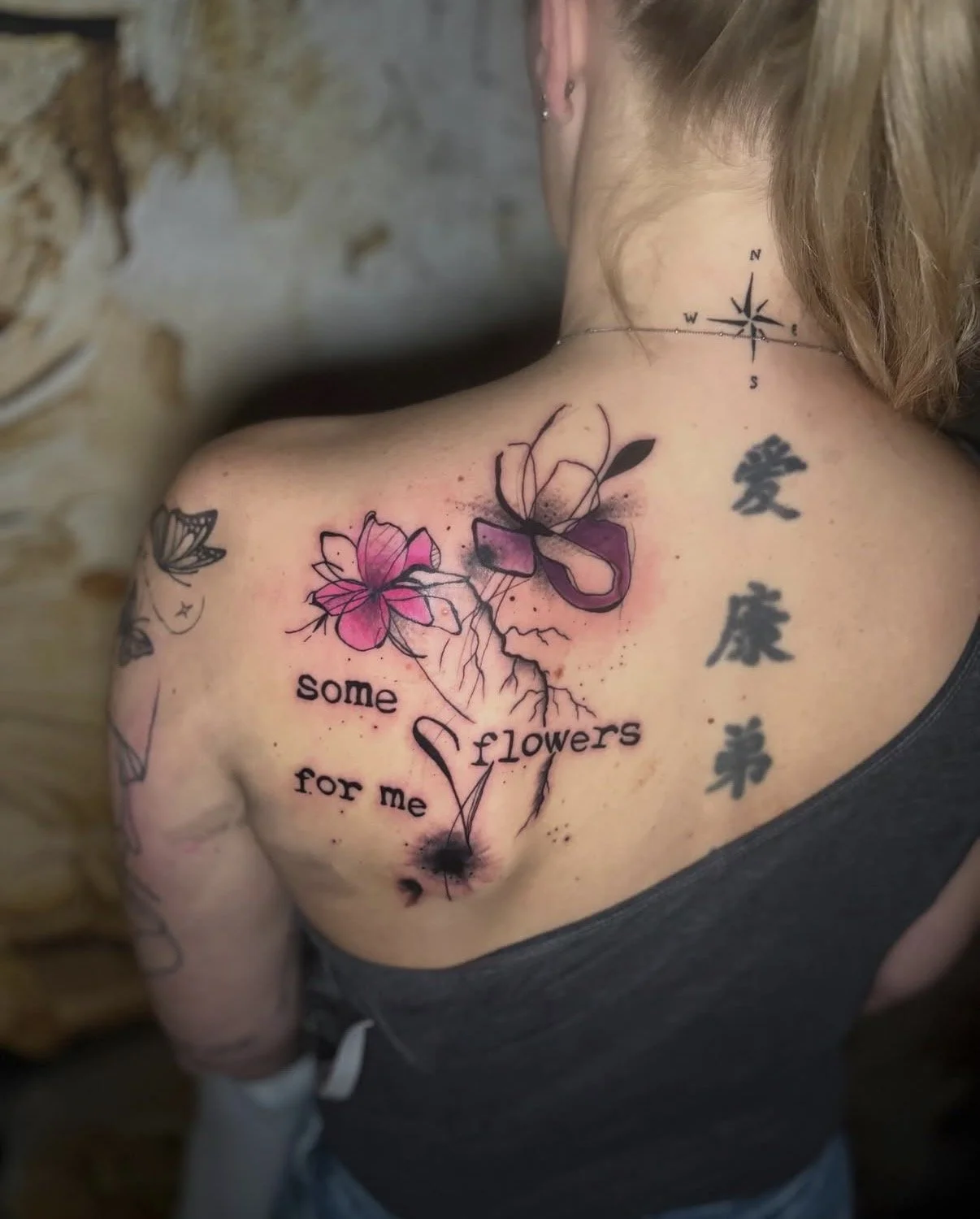 Tattoo auf der Schulter einer Frau mit pinken und schwarzen Blumen, einem Herz, einem Puzzle-Teil, chinesischen Schriftzeichen und dem Text 'some flowers for me'
