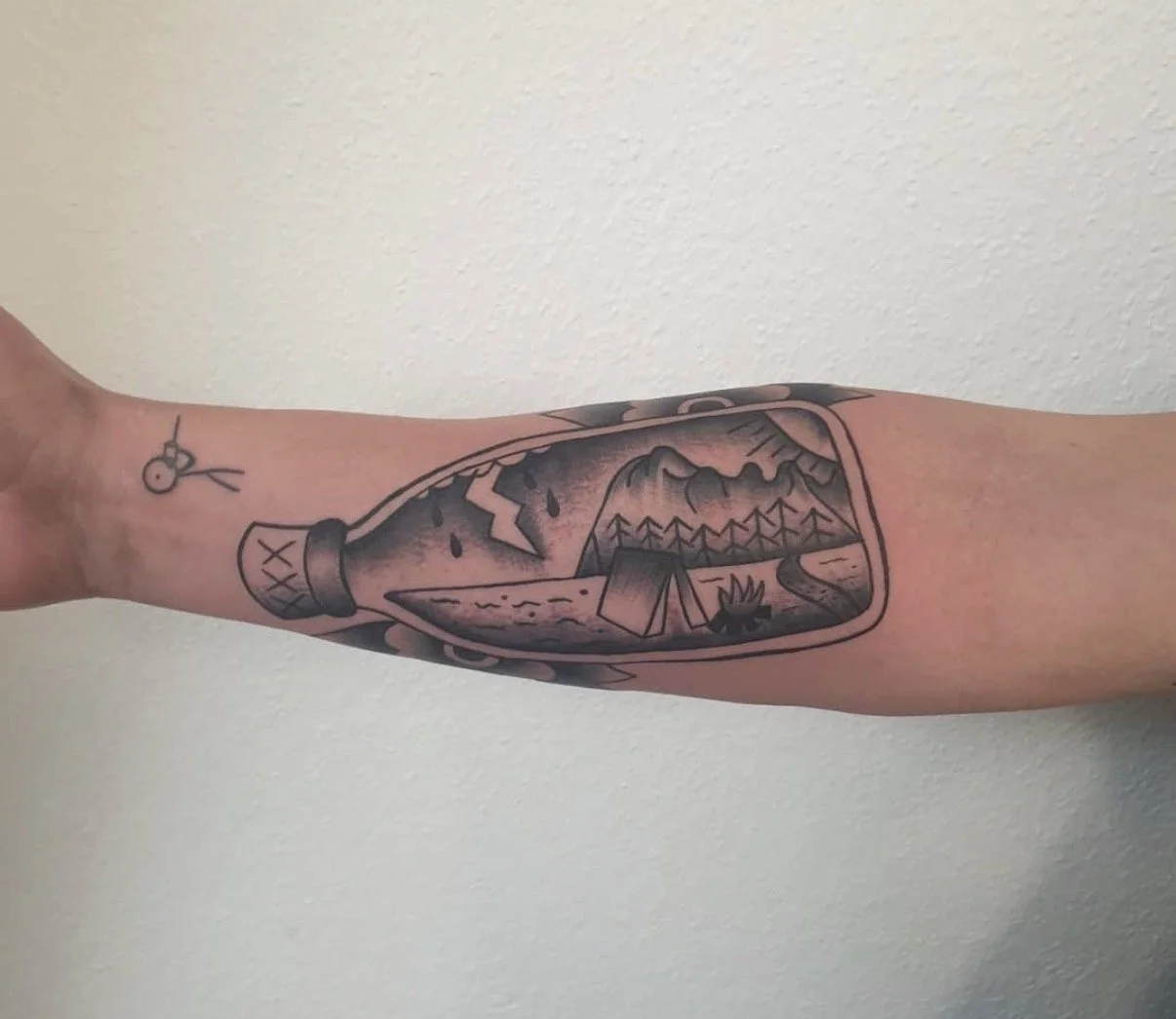 Tattoo auf dem Unterarm, zeigt eine Landschaft mit Bergen, Bäumen, Fluss und einem Zeltdach, eingeschlossen in eine Flasche.