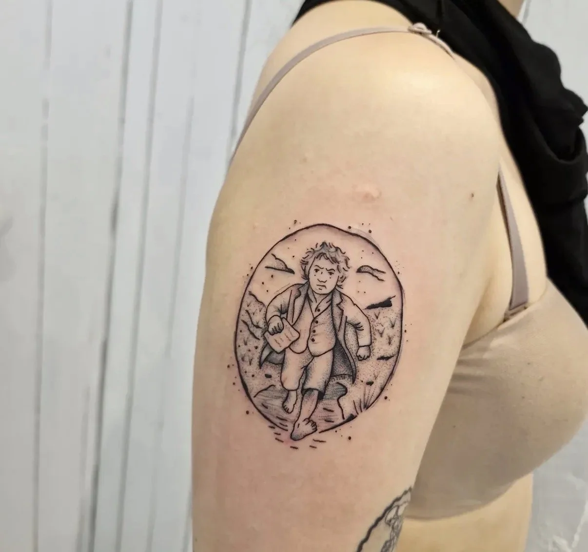 Schwarz-weiße Tattoo auf der Oberarm einer Person, zeigt eine Figur im historischen Kleid, die mitten im Schritt läuft, mit einem Buch in der Hand, in einer Umgebung mit Bergen und Vögeln im Himmel, within a circular Rahmen.