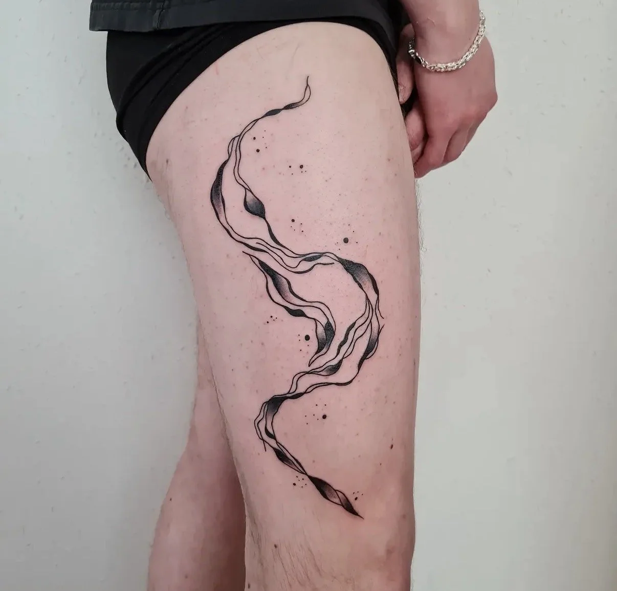 Schwarzes und graues Tattoo in abstrakter, wellenförmiger Linienführung auf dem Oberschenkel einer Person, mit kleinen schwarzen Spritzern und Dotwork, Person trägt schwarzen Hosenbund und eine Silberkette am Handgelenk.