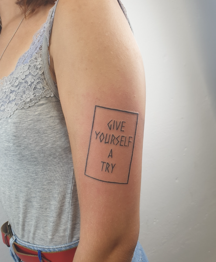 Tattoo auf Oberarm mit Text: 'GIVE YOURSELF A TRY', umrahmt von einem rechteckigen Rahmen.