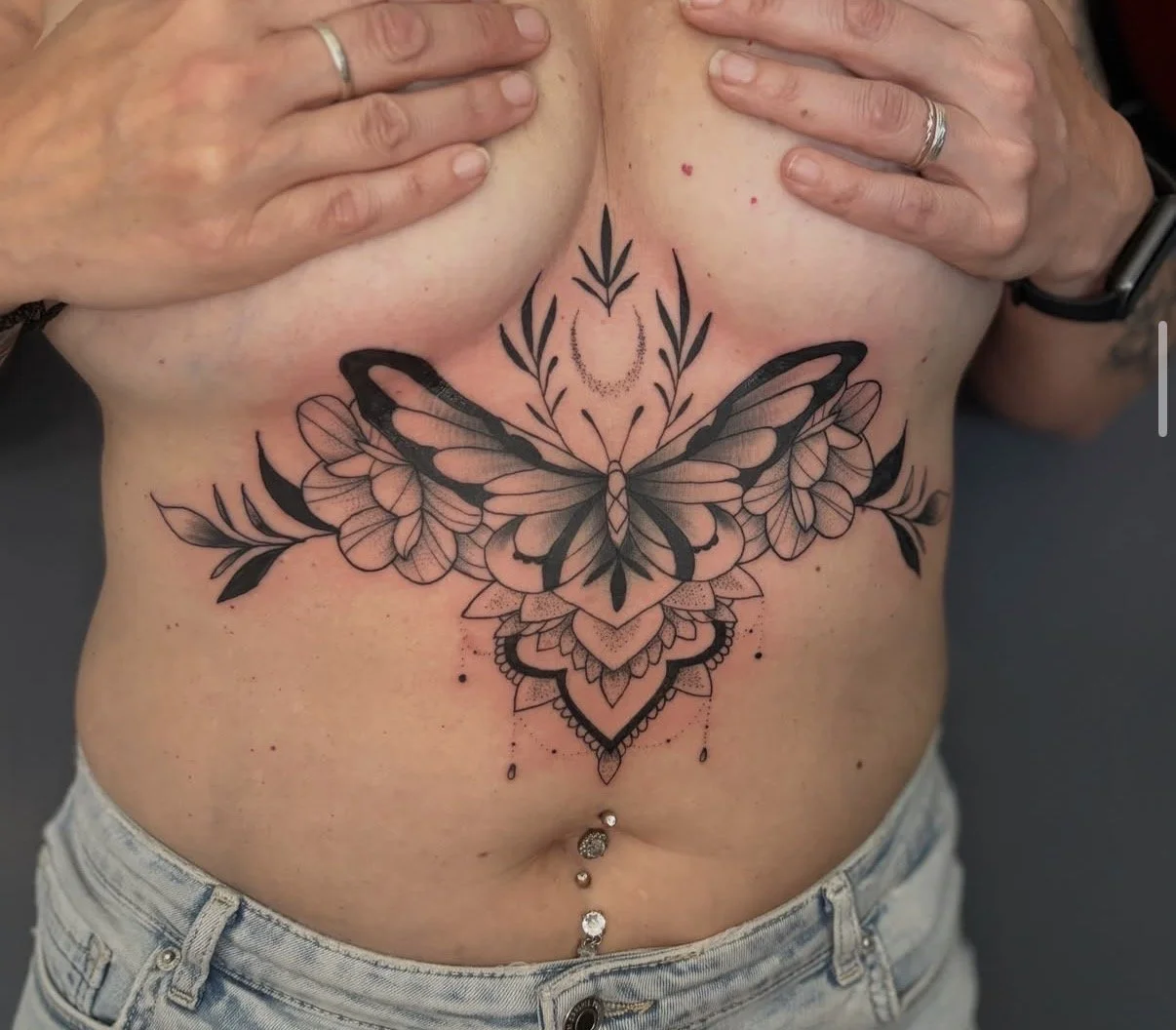 Schmetterling-Tattoo auf dem Bauch einer Person, mit Blumen, Blättern, einem Halbmond und dekorativen Elementen in schwarz.