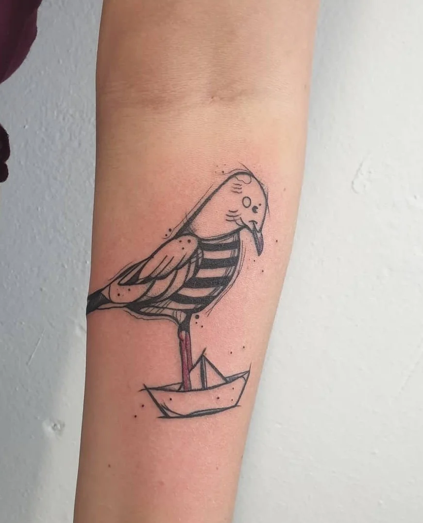 Tattoo eines Vogels auf dem Unterarm, stilisiert mit Linien, in einem Papierboot stehend, das auf der Haut sichtbar ist.