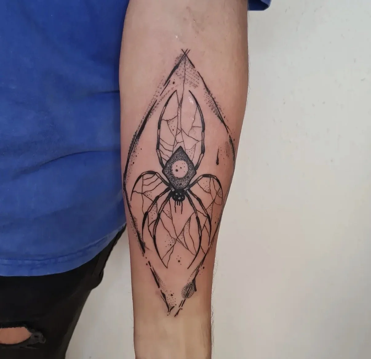 Tattoo eines Spinnenwebers auf dem Unterarm, mit einem Spinnenmotiv in der Mitte, in schwarzer Tinte mit geometrischen und punktierten Details.