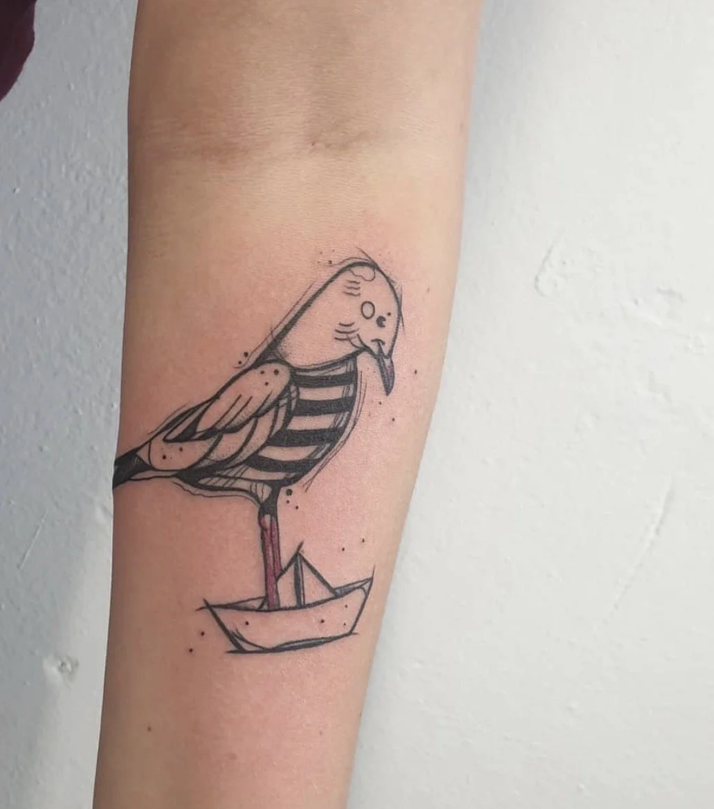 Tattoo eines Vogels mit einem Papierboot als Körper auf dem Unterarm.