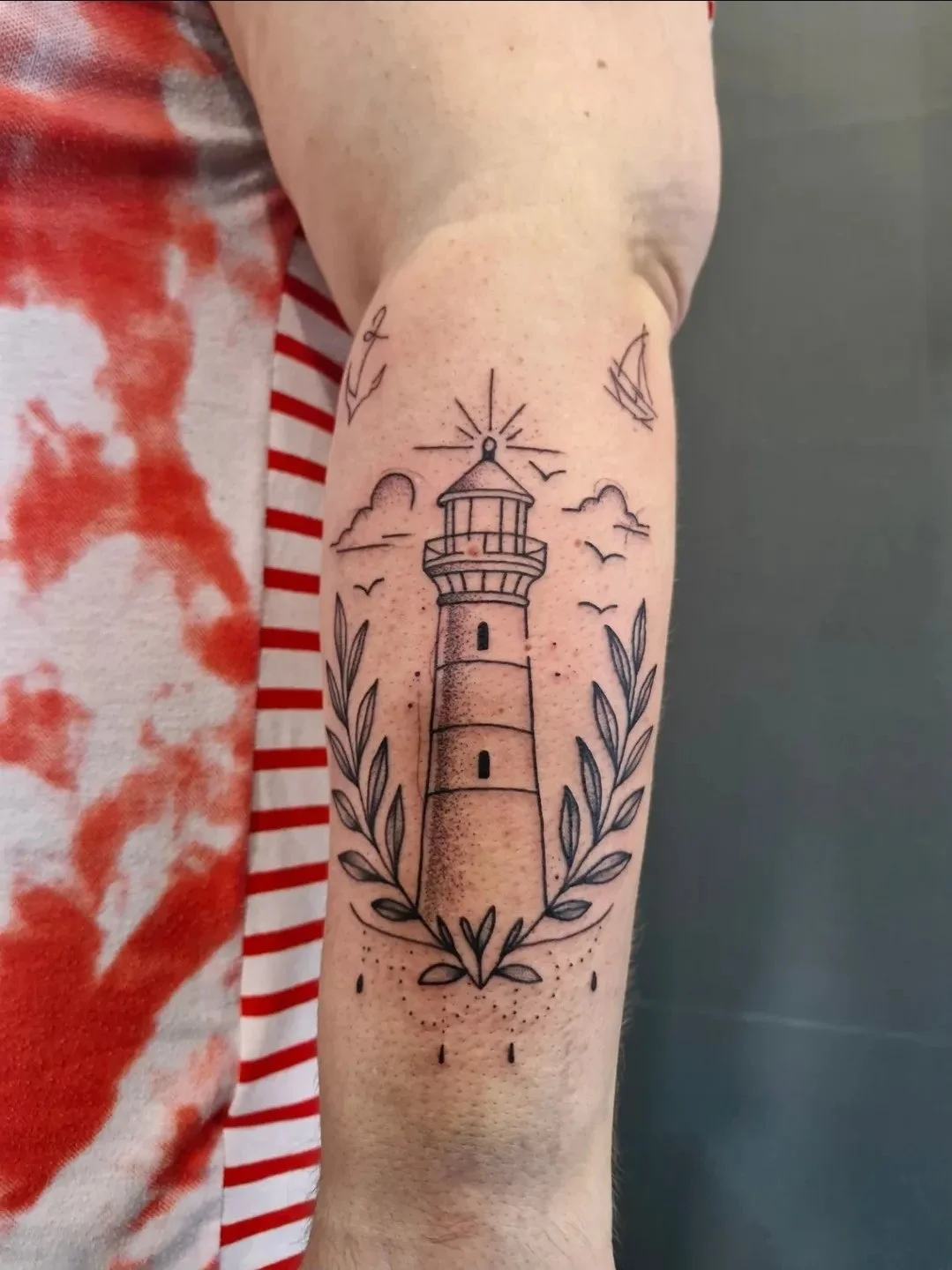 Tattoo eines Leuchtturms mit Sonnenstrahlen, Vögeln und Blättern auf dem Oberarm.