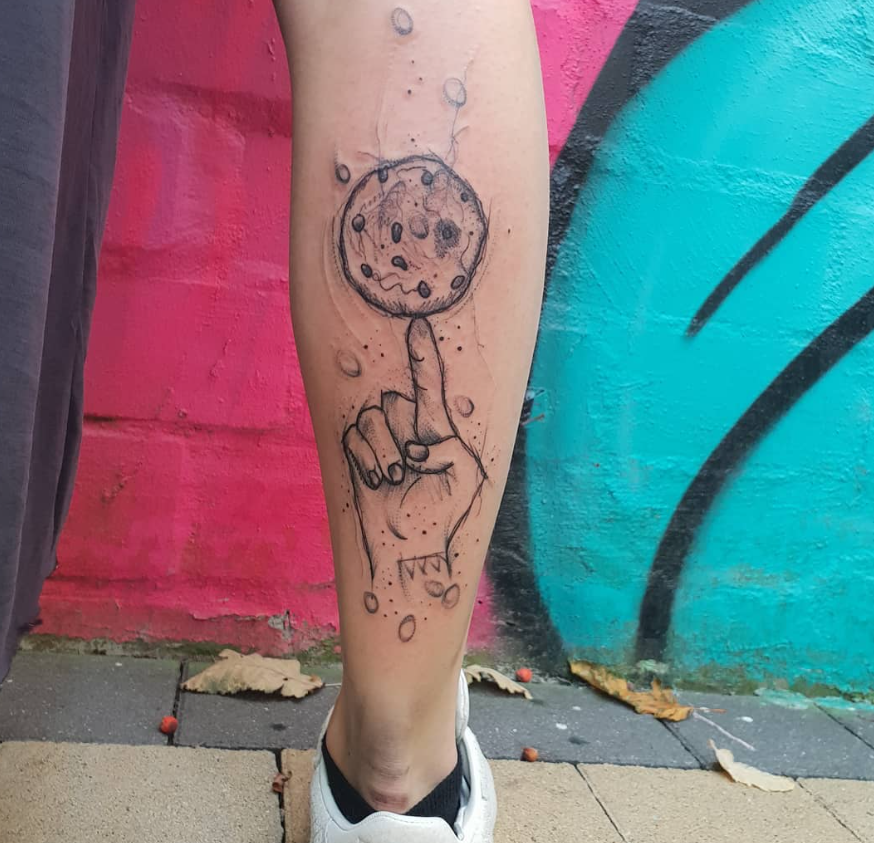 Tattoo einer Hand, die einen Finger mit einem Keks auf einem Arm hält, vor einem bunten Wandgemälde.