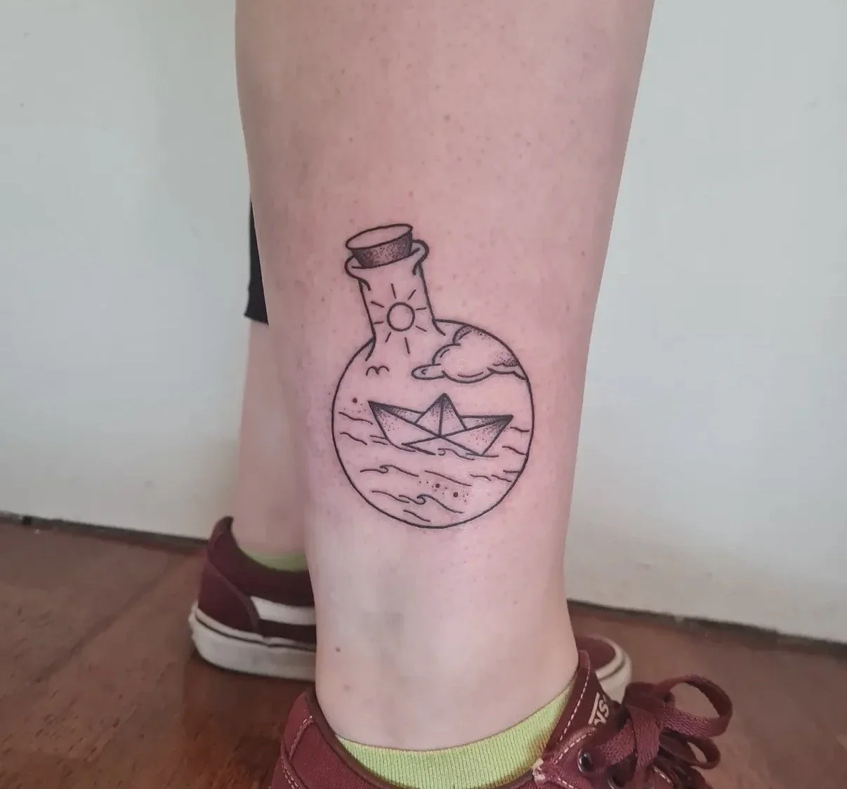 Tattoo eines Flakons mit einer Sonne, Wolken, Meer und einem Papierboot darin.