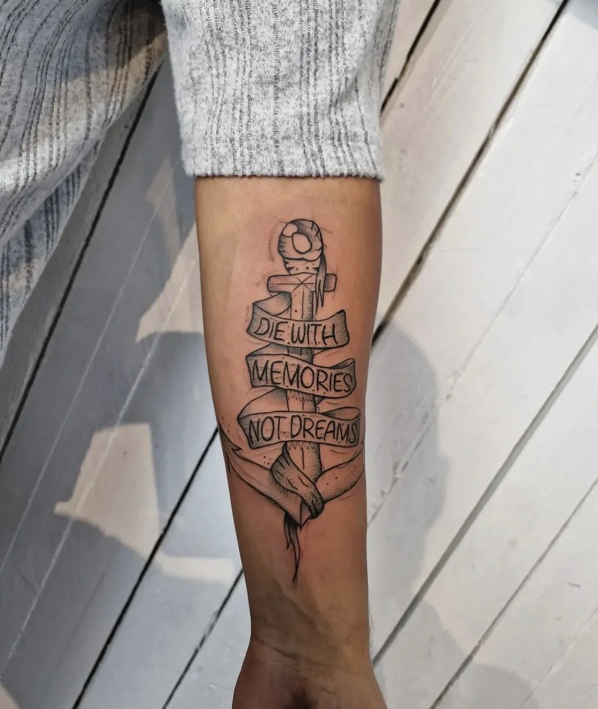 Tattoo eines Ankers mit einem Spruchband, das die Worte 'DIE WITH MEMORIES NOT DREAMS' trägt, auf einem Unterarm.