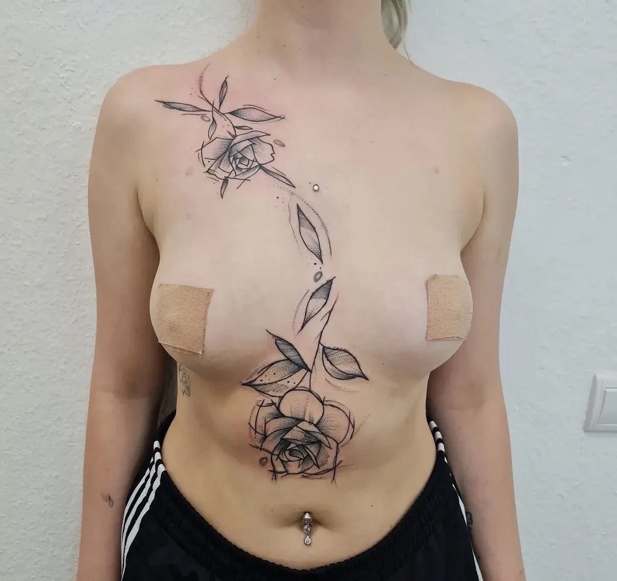 Eine Frau mit neuen Tattoos auf der Brust, die Rosen und Blätter zeigen, sowie ein Bauchnabelpiercing, die mit Pflastern bedeckt sind.