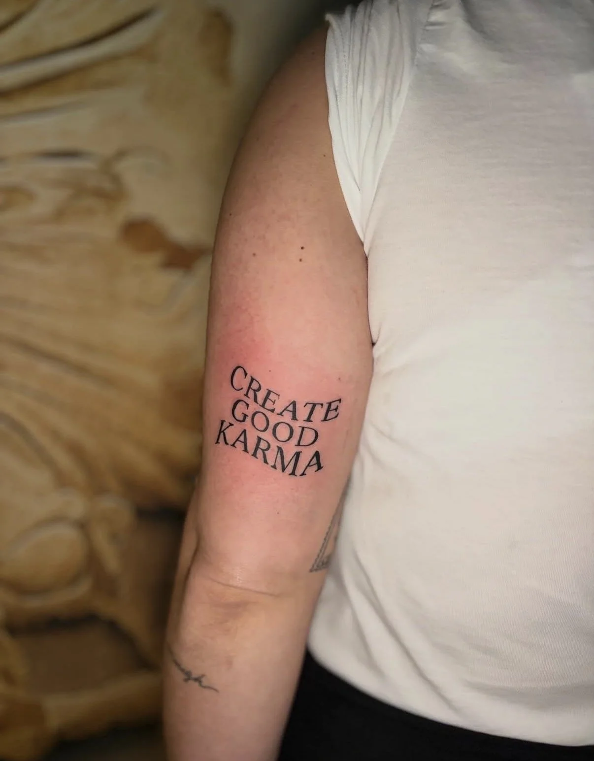 Tattoo auf dem Oberarm mit dem Text "Create Good Karma" in schwarz. Person trägt ein weißes T-Shirt, Hintergrund besteht aus Holz.