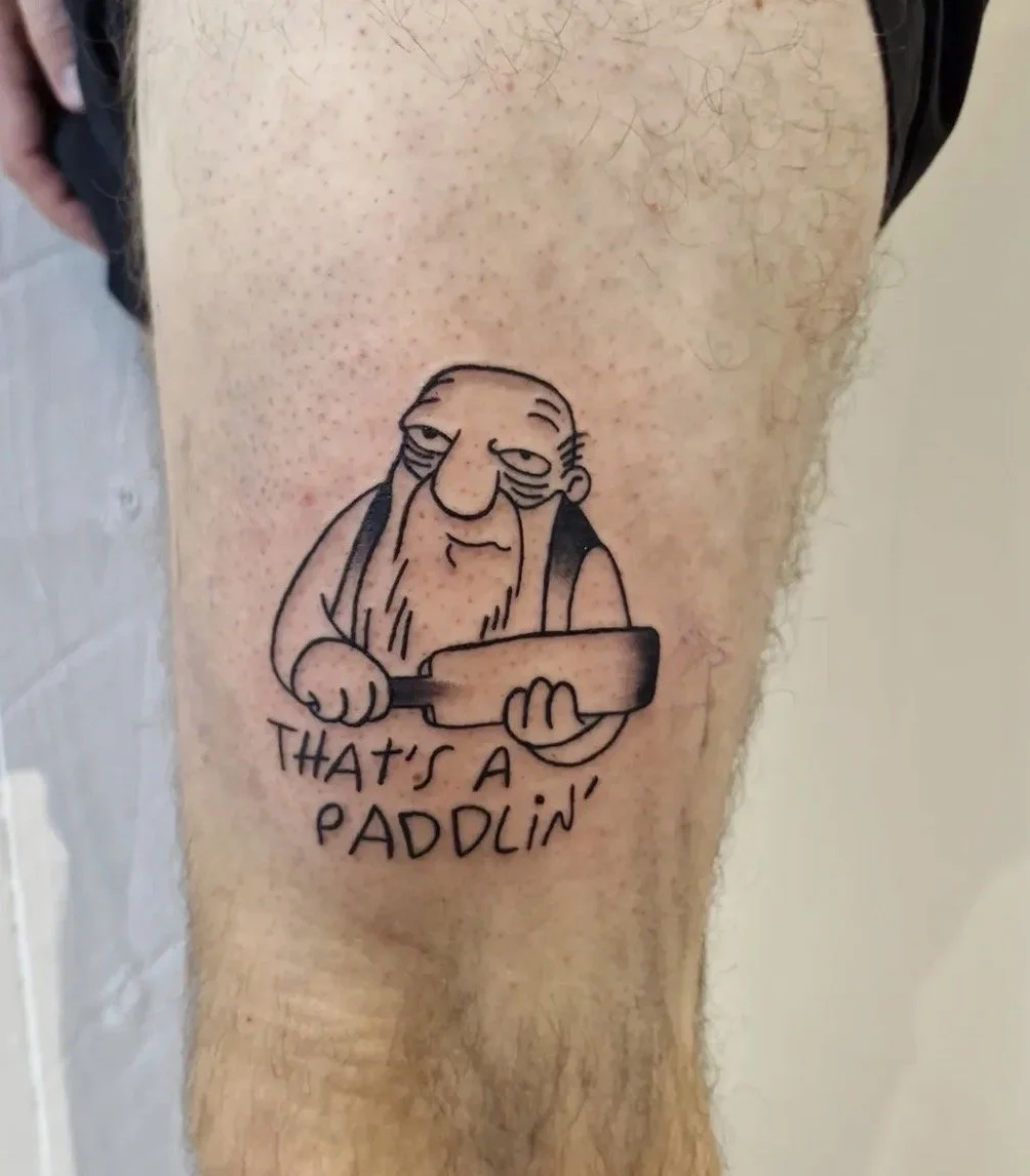Tattoo eines alten Mannes mit langem Bart, der ein Holzstück hält, und der Text 'That's a paddlin'' darunter.