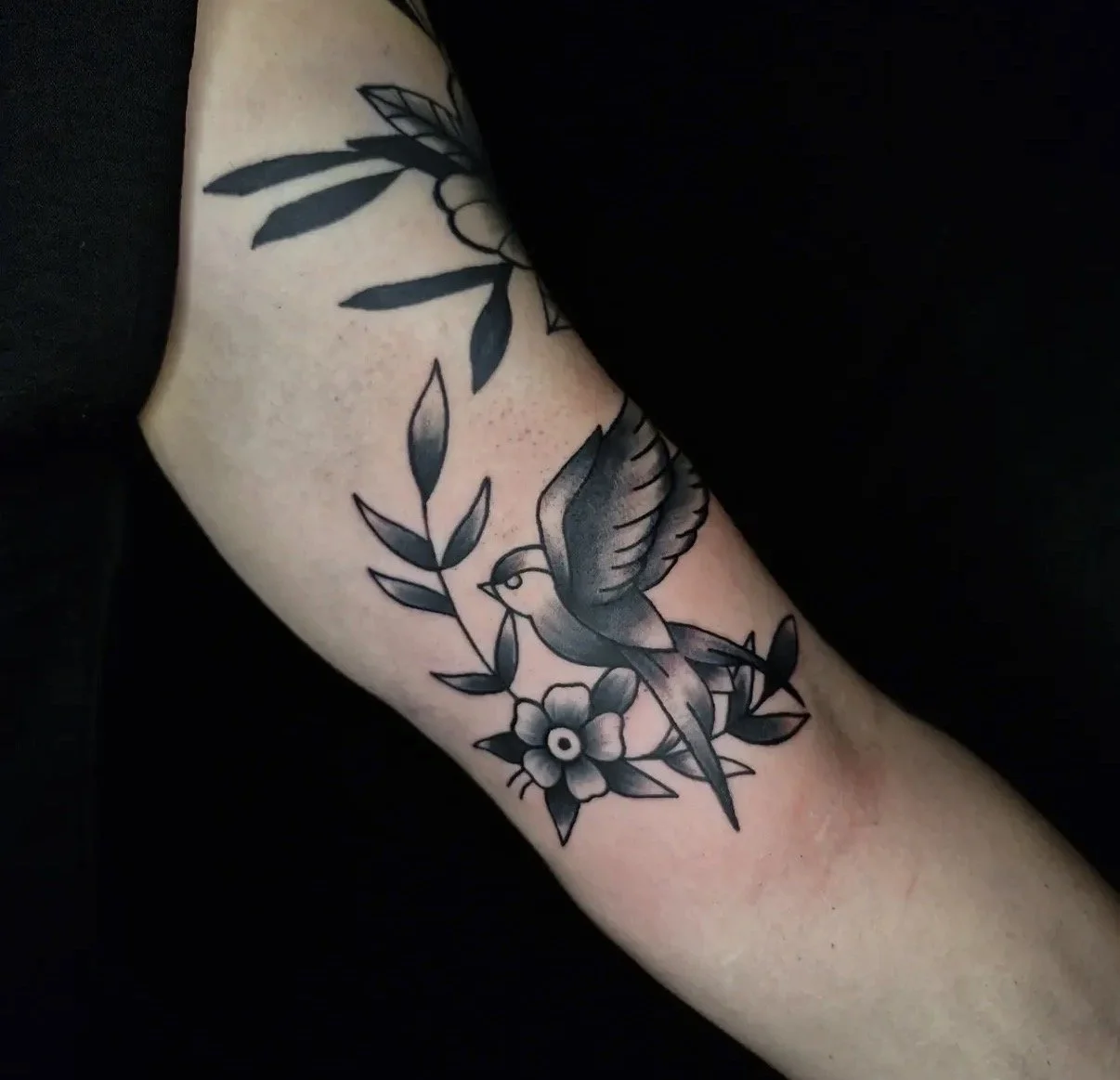Schwarz-weiß Tätowierung eines Vogels, der auf einer Blume sitzt, umgeben von Blättern auf dem Arm.