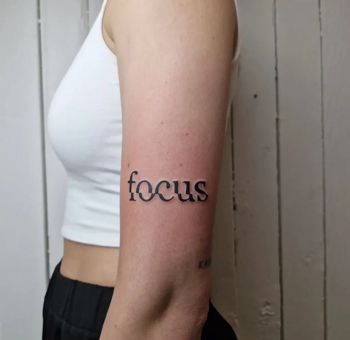 Nahaufnahme eines Arms mit einer Tätowierung, auf der das Wort 'focus' steht, vor einer weißen Holzwand.