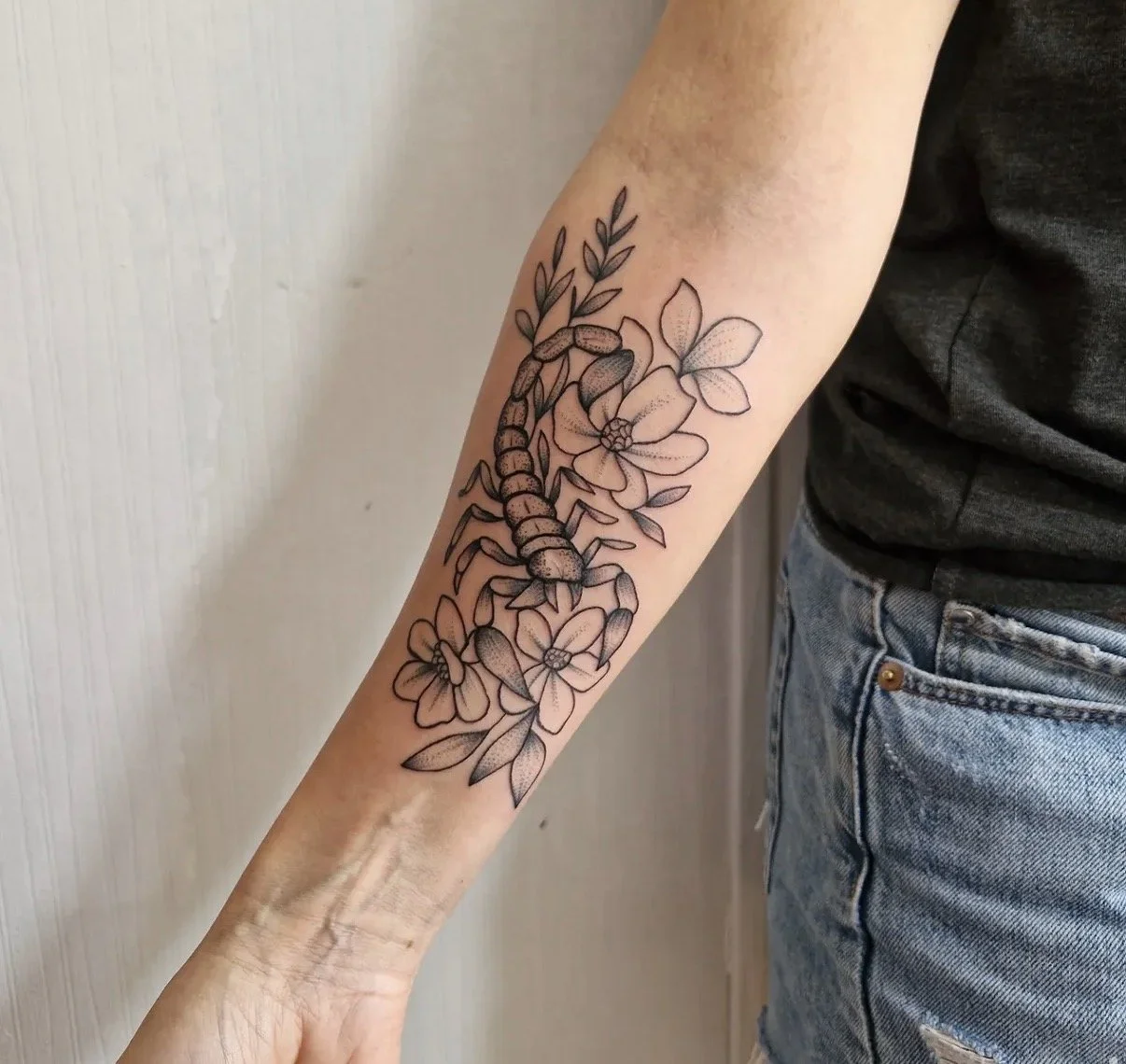 Schwarzer Tattoo eines Skorpions, umgeben von Blumen und Blättern, auf dem Unterarm einer Person.