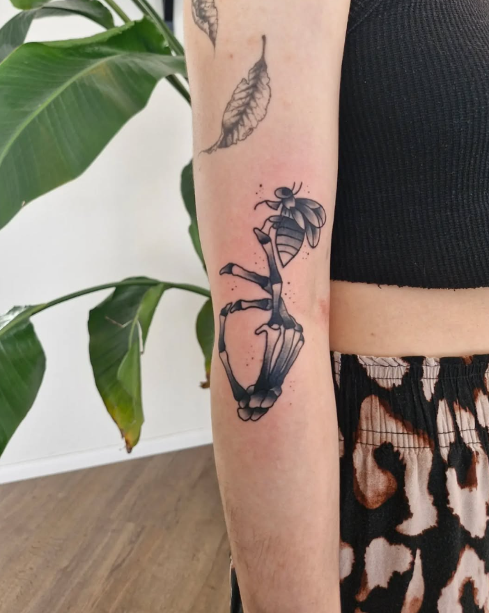 Tattoo einer Biene und eines toten Insekts auf dem Unterarm einer Person, im Hintergrund grüne Pflanzen.