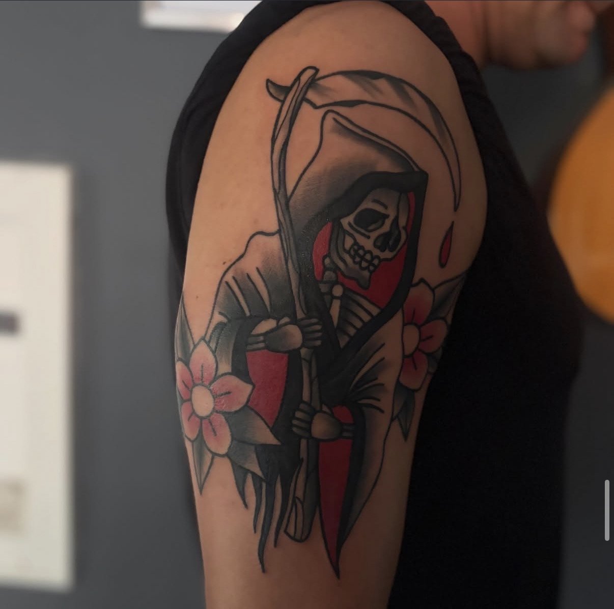 Tattoo eines Skeletts in einem dunklen Hoodie, umgeben von Kirschblüten auf dem Oberarm eines Menschen.