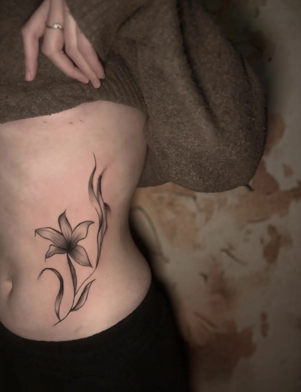 Schwarze Tattoo-Blume auf Körperseite, person trägt braunen Pullover.