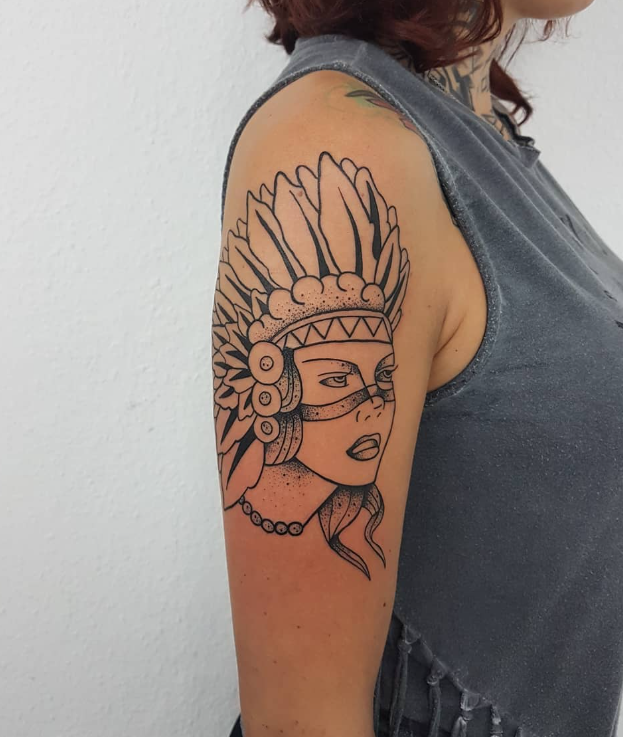 Tattoo einer weiblichen Figur mit Indianer-Headdress auf dem Oberarm einer Person, die grau ärmelloses Top trägt.