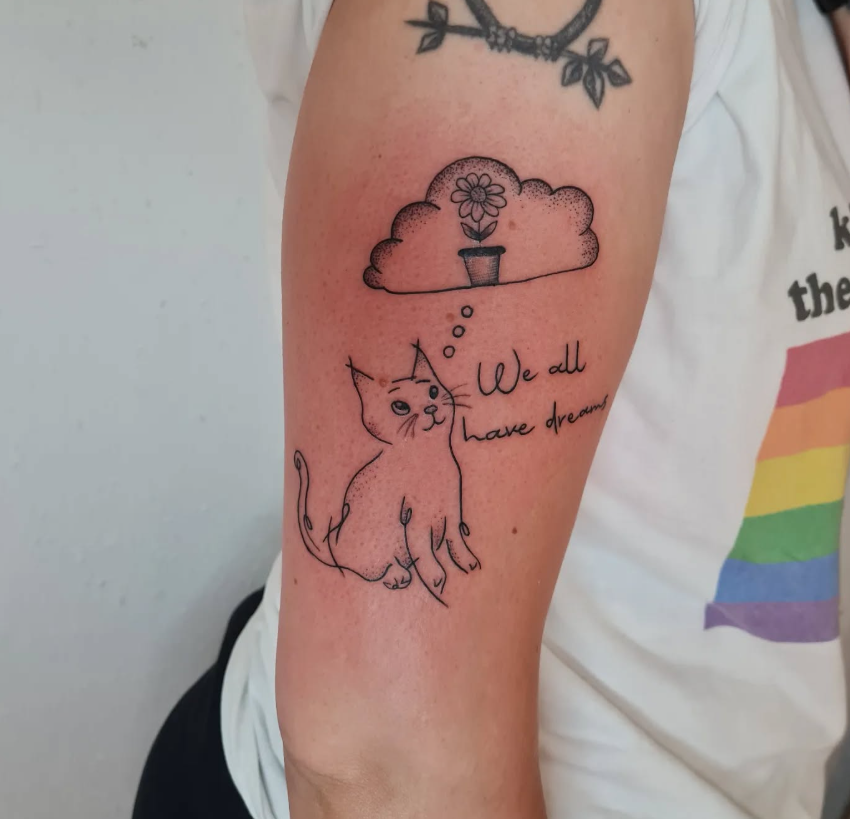Tattoo eines sitzenden Katers mit Gedankenblase, die eine Blumenvase enthält, sowie der Text 'We all have dreams'.