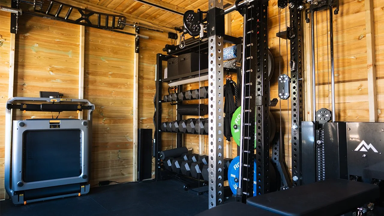 Home gym with wooden walls, featuring a squat rack, weight plates, dumbbells, a foldable treadmill, and various workout accessories.