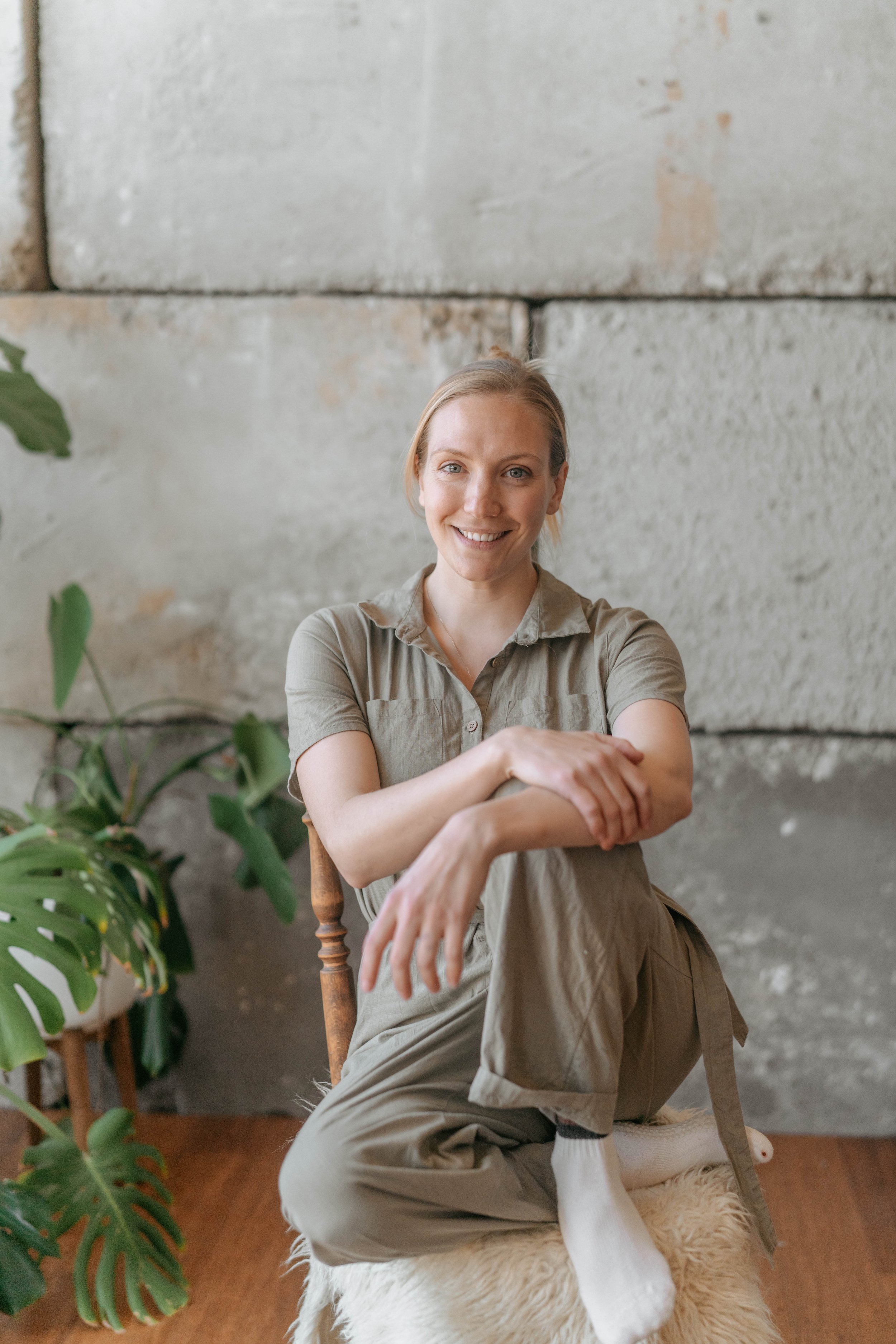 A woman with blonde hair in a bun, smiling, sitting on a fur-covered stool in front of a gray stone wall, wearing a beige shirt and khaki pants with socks, with green plants nearby.