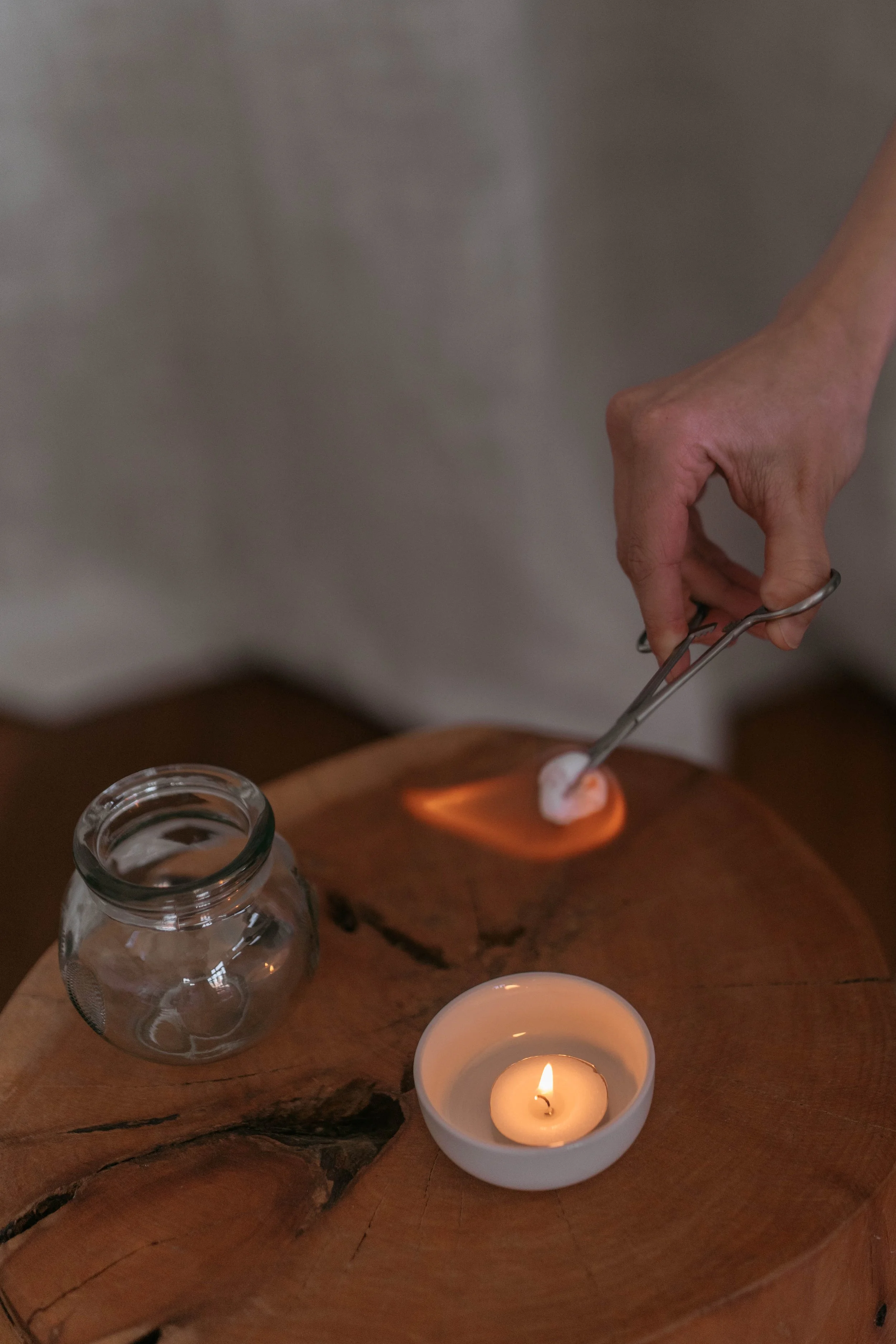 Hand of a person holding a dental impression tool above a lit candle on a wooden surface, with a jar and a cup nearby, in a dimly lit environment.