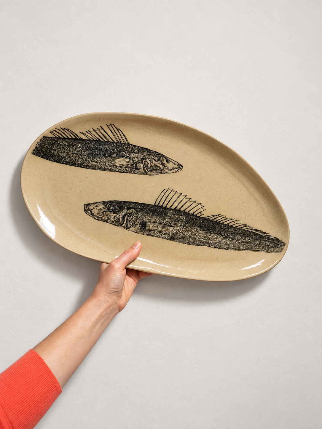 Person holding a beige oval plate with black printed fish illustrations on a white background.