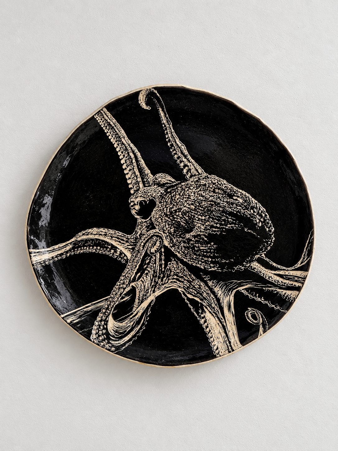 A black decorative plate with an illustration of an octopus in beige, featuring detailed tentacles and head, on a white surface.