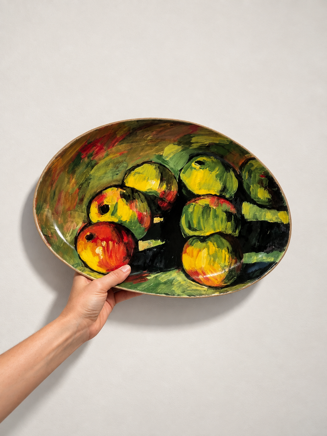 Colorful oval bowl with a painted still life of apples held against a light gray wall.