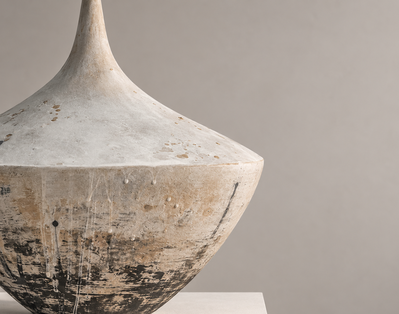 Close-up of a weathered, beige, and black ceramic vase with a narrow neck, positioned on a white pedestal against a plain gray background.