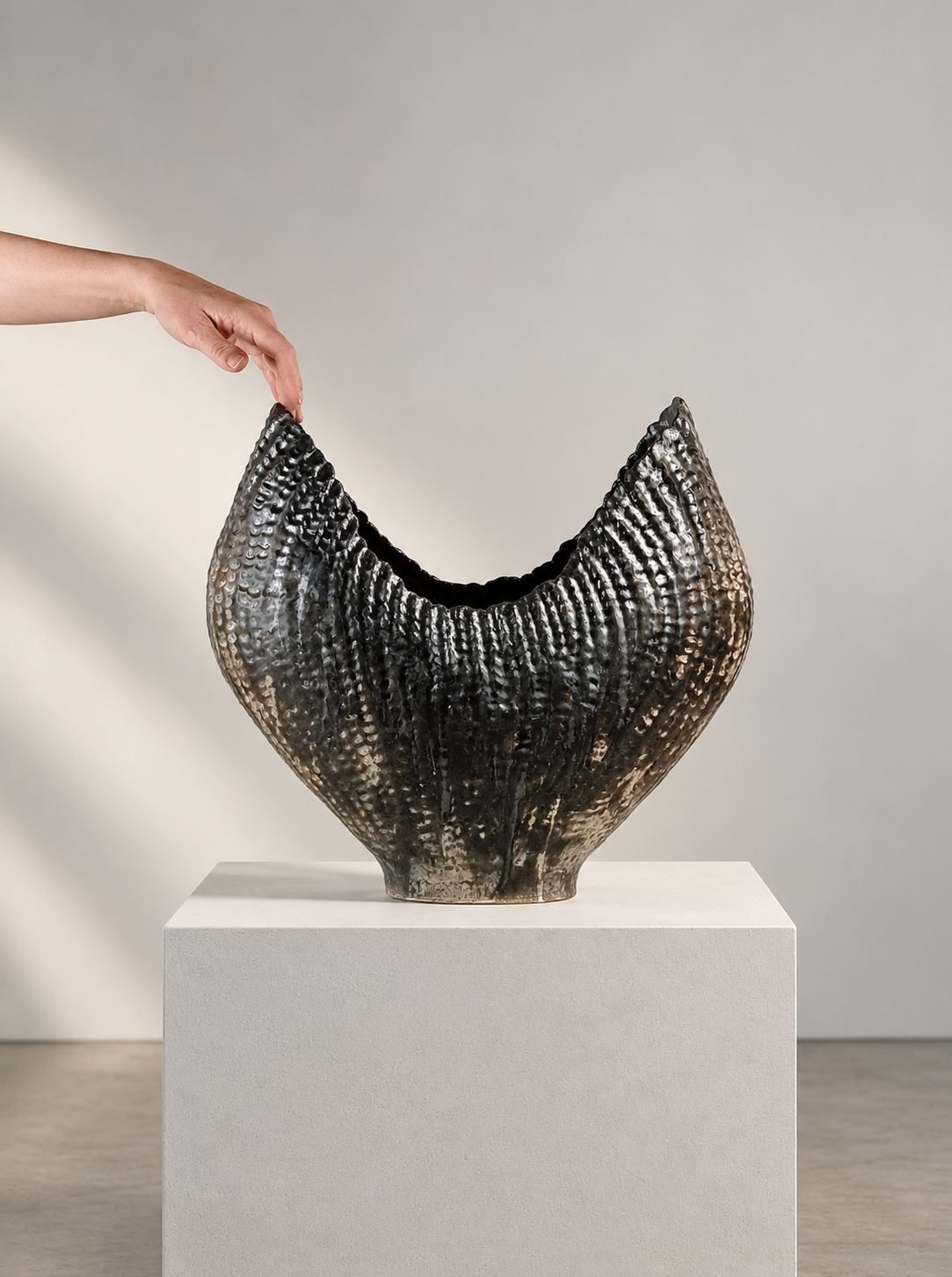 A large, textured black and white vessel on a white pedestal with a person's hand reaching to touch it.