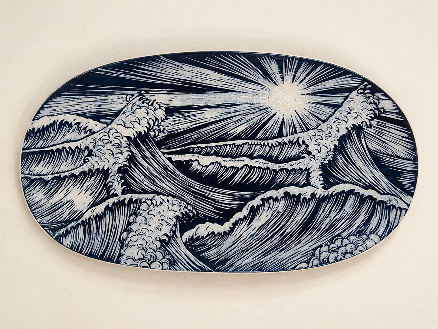 An oval ceramic plate with a blue and white design of stylized crashing waves and a bright sun or moon in the sky.