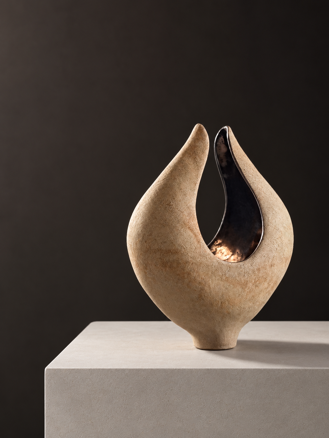 A modern sculpture with a curved, teardrop shape, made of a beige textured material with a shiny metallic interior, displayed on a white pedestal against a dark background.