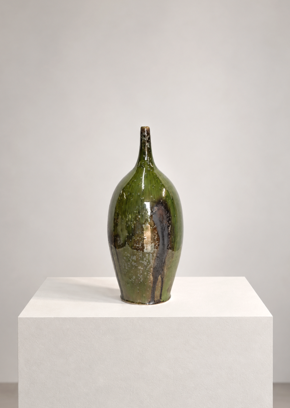 A tall, green and brown ceramic vase with a narrow neck, displayed on a white pedestal against a plain white background.