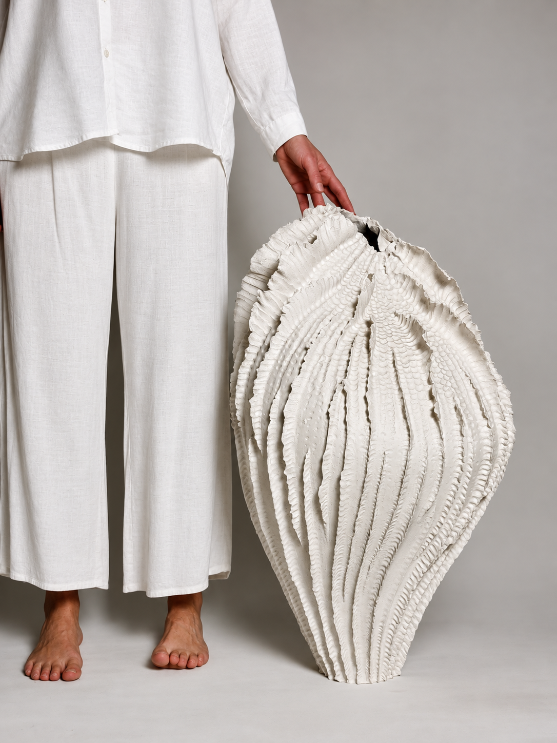 Person in white linen clothing standing barefoot next to a large, white, textured seashell vessel on a neutral background.