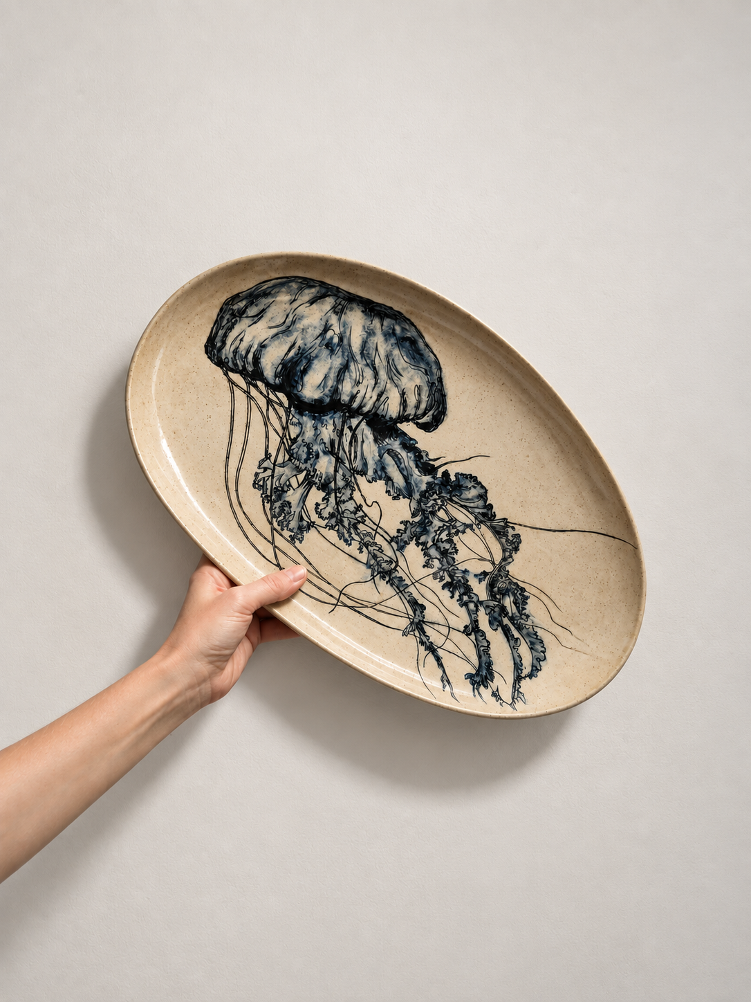 A hand holding an oval beige ceramic plate featuring a blue illustration of a jellyfish with long tentacles.