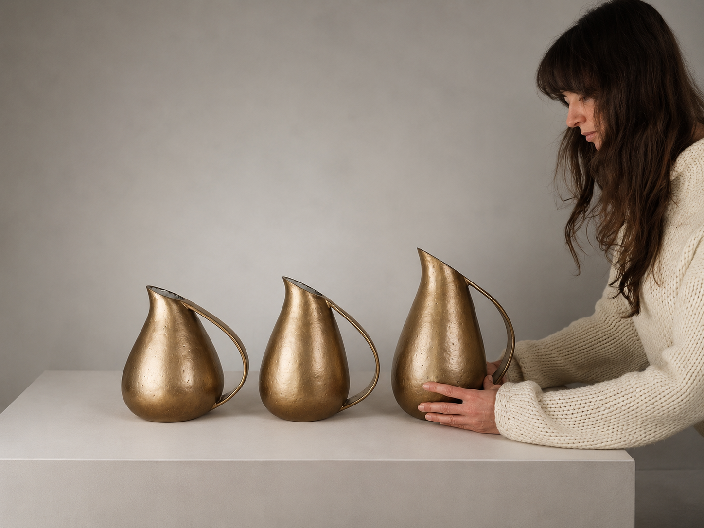 Woman in cream sweater arranging three gold decorative pitchers on a white table against a neutral background.