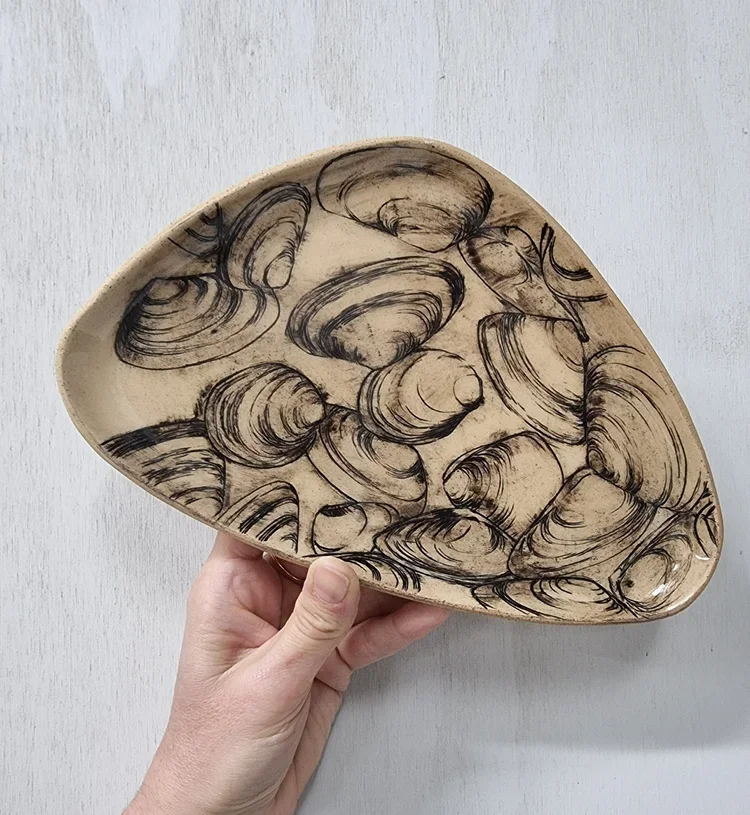 Hand holding a ceramic plate with sketches of seashells drawn in black ink.