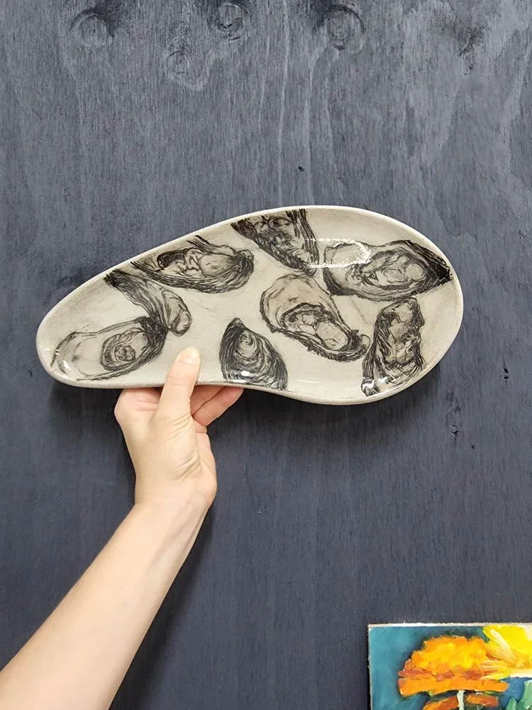 Hand holding a ceramic dish with black line drawings of oyster shells on a dark surface.