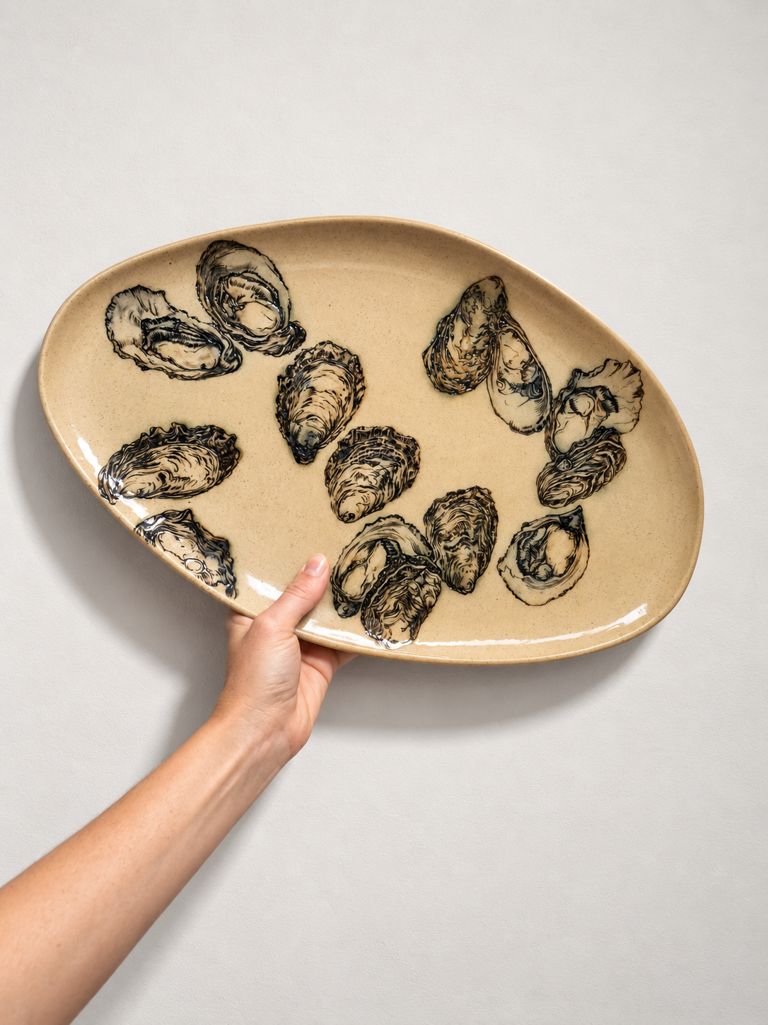 Hand holding a ceramic serving platter with painted oyster shells on a light background.