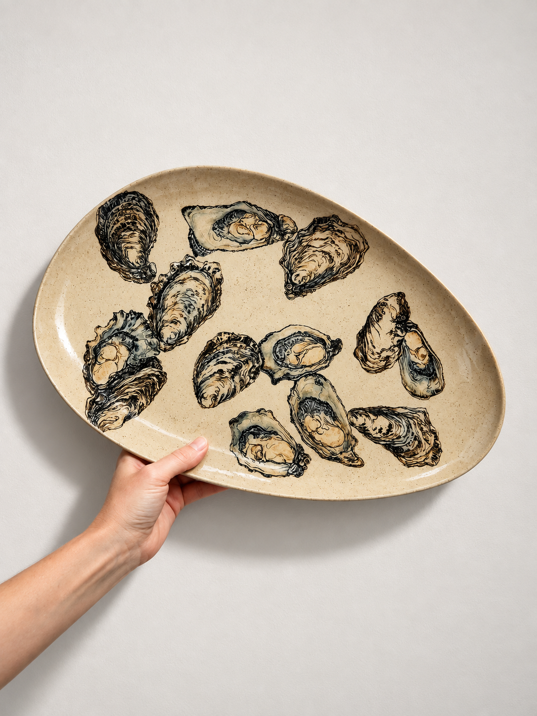 A hand holding a beige oval ceramic plate with a black shell pattern, placed on a plain white surface.