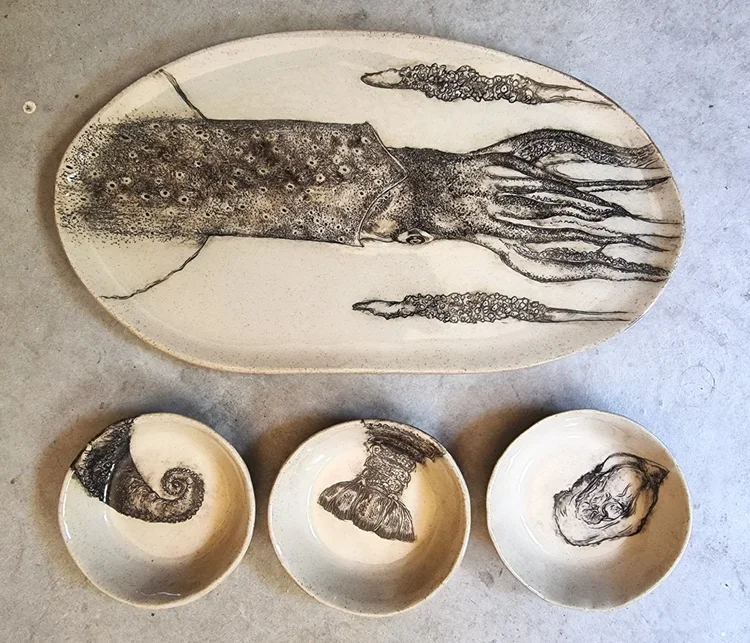 Ceramic dishware set featuring sketches of a squid, a spiral shell, a mushroom, and a stone or fragment, all decorated in black ink on a cream-colored background.