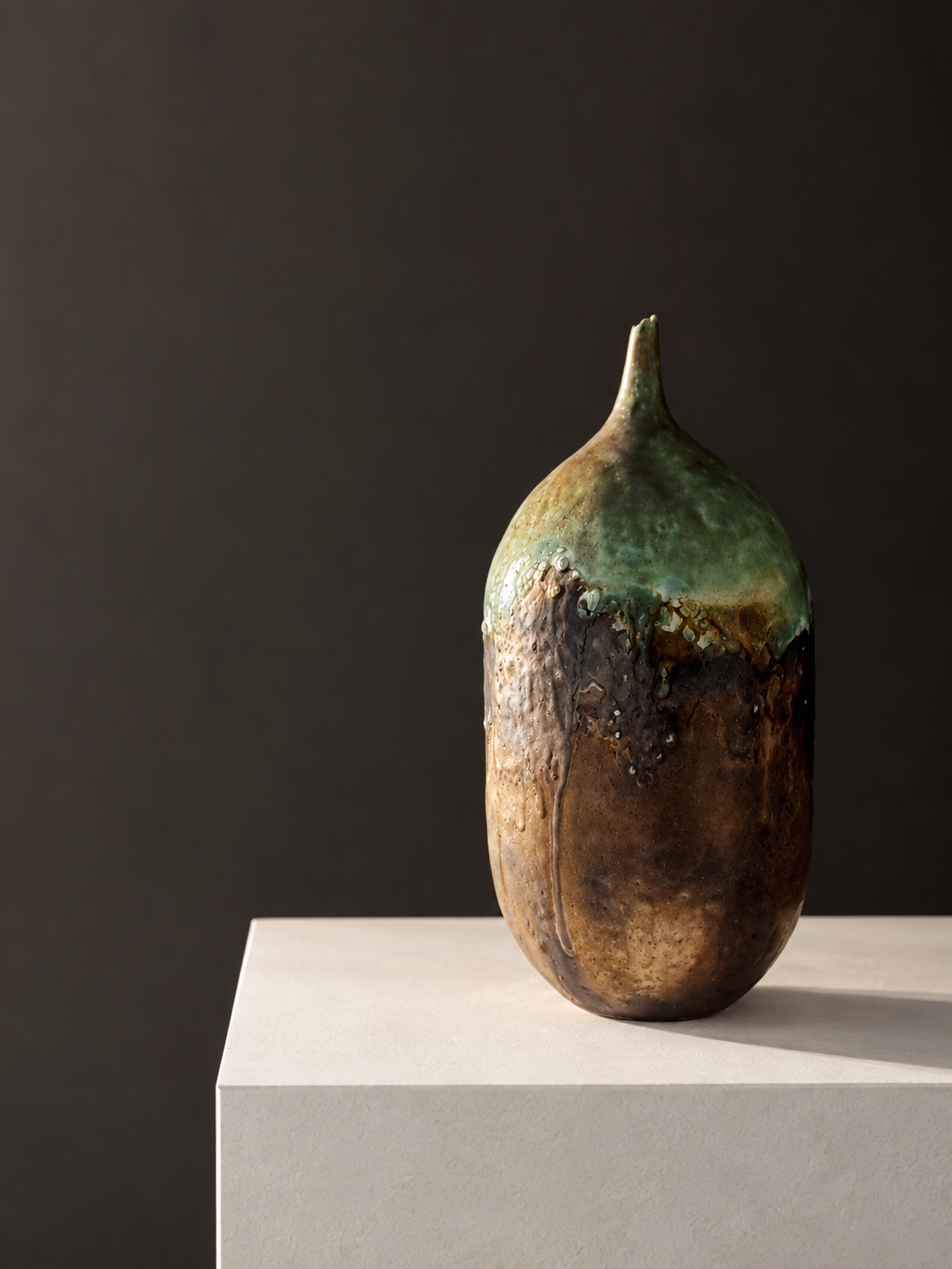 Ceramic sculpture resembling a stylized seed or pod with earthy tones of brown, green, and beige, placed on a white pedestal against a dark background.