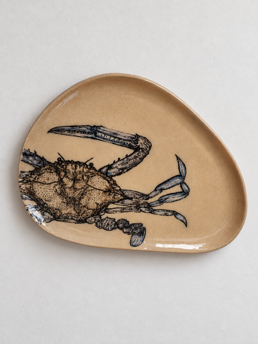 A beige, irregularly shaped ceramic plate with a detailed black and blue illustration of a crab.