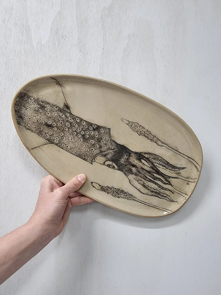 Oval ceramic plate with a black ink drawing of a squid on it, held by a person's hand against a white wall.