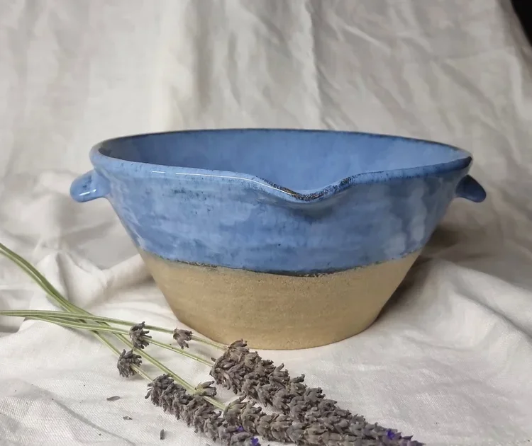 Ceramic bowl with blue glazed top half and unglazed bottom half, placed on white fabric with dried lavender flowers in front.