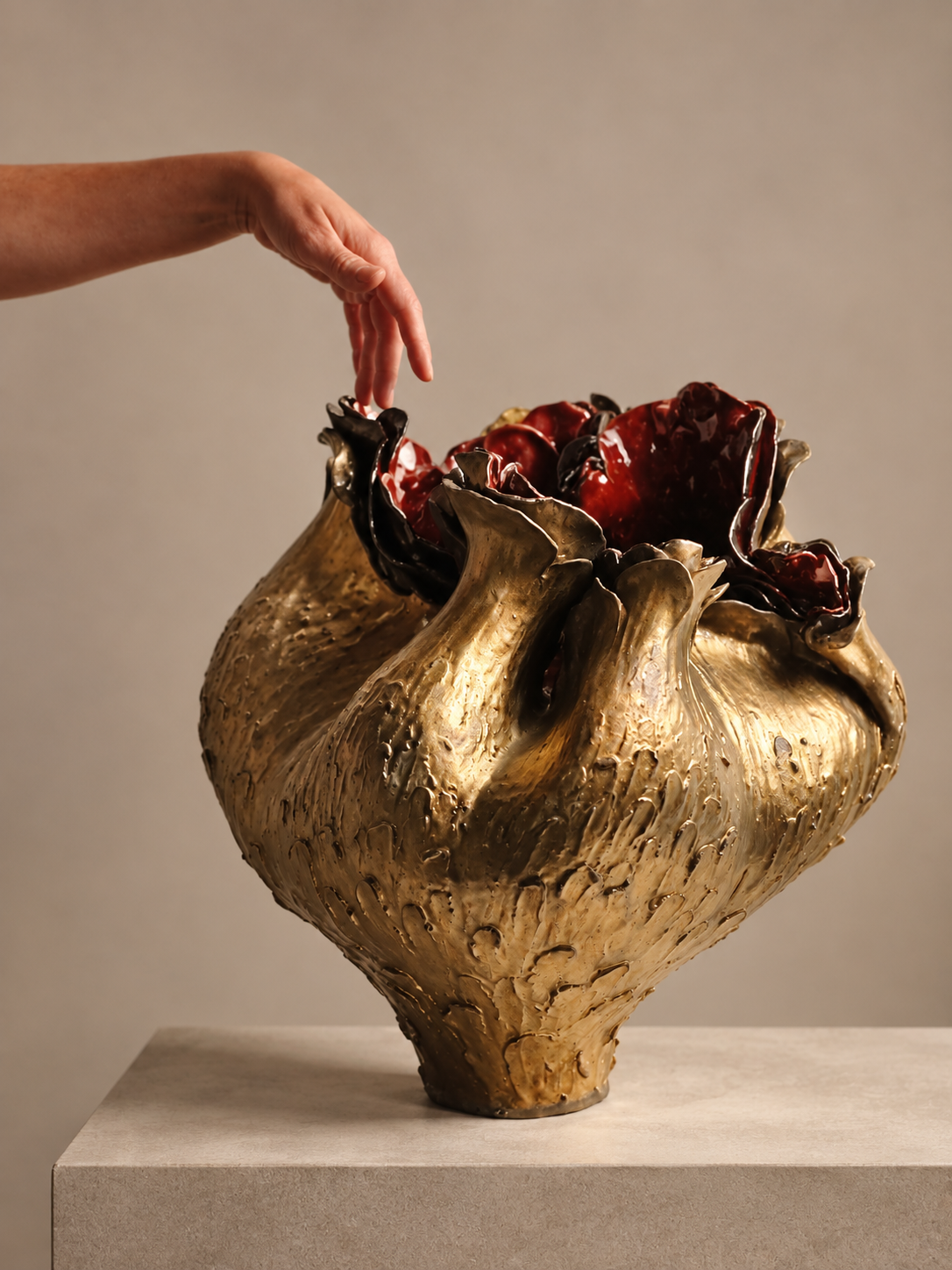 Close-up of a hand reaching towards a large, artistic ceramic vase with an irregular, organic shape and a textured beige exterior. The top of the vase is open, revealing dark red and black glossy interior details, and it is placed on a plain pedestal against a neutral background.