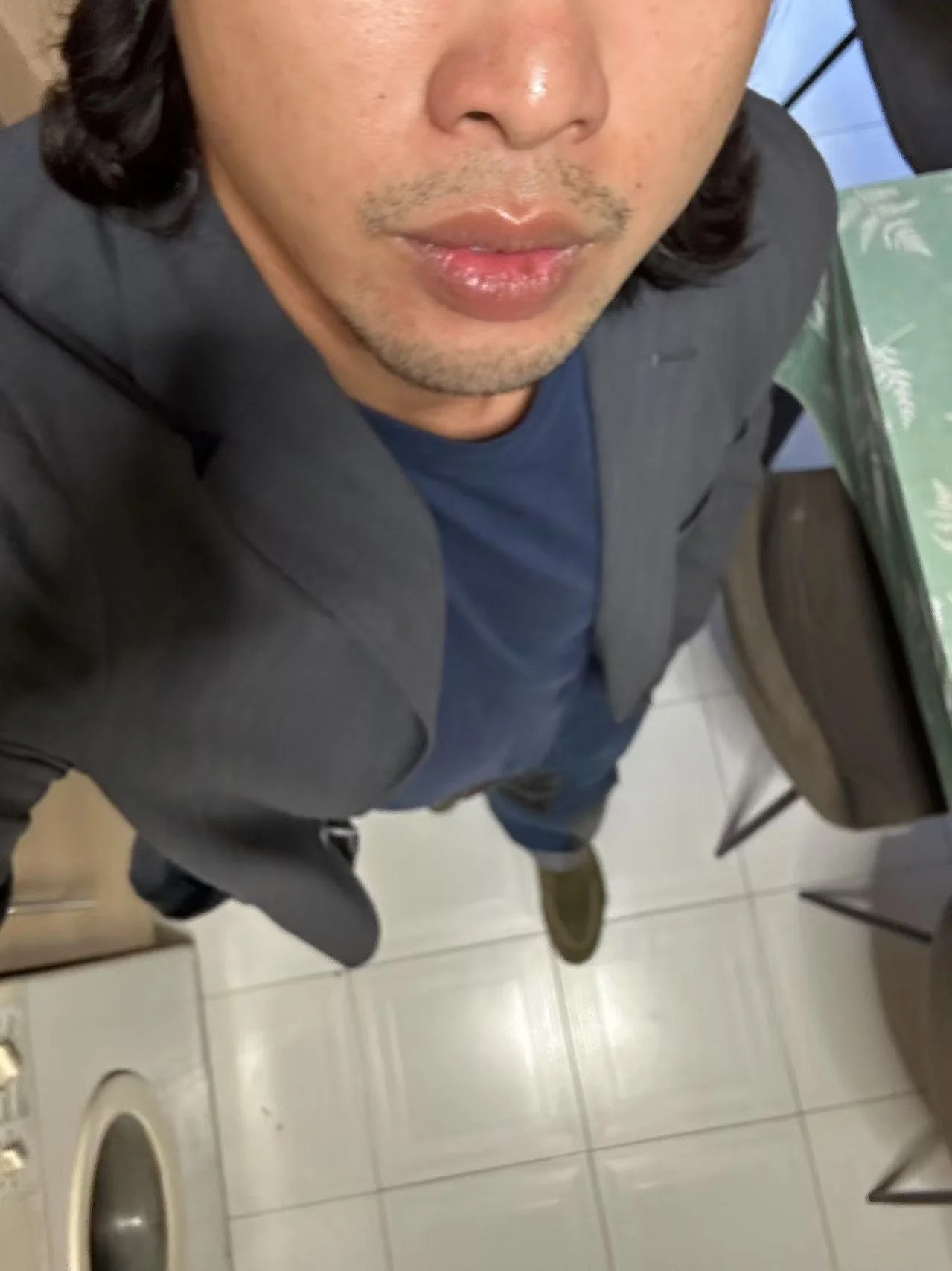 A man is standing in a bathroom taking a selfie from a high angle showing his face, gray blazer, blue shirt, and shoes. The bathroom has white tiles, a toiletry container, a green tablecloth with white patterns, and a chair.