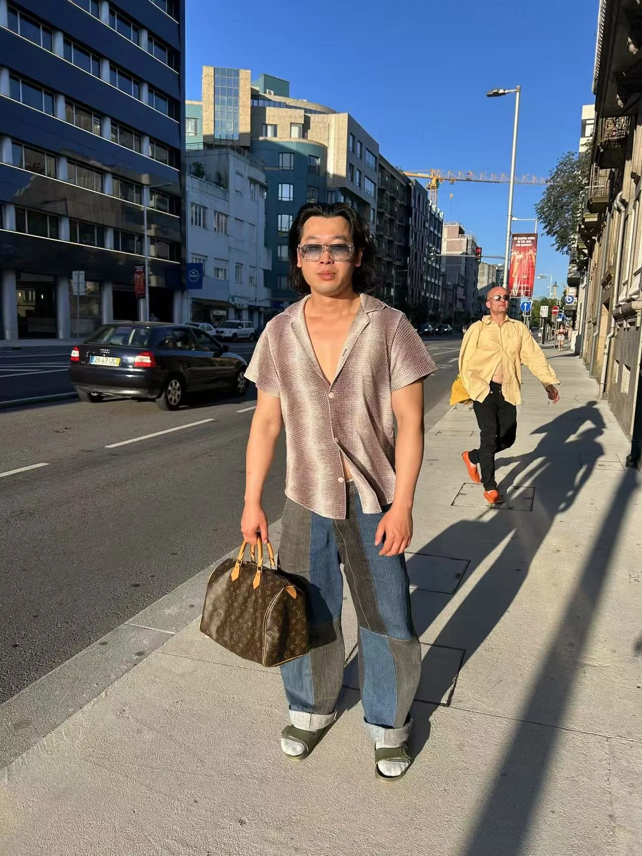 A stylish person with long dark hair, wearing sunglasses, a shiny beige shirt, baggy plaid jeans, and sneakers, stands on a city sidewalk holding a Louis Vuitton handbag. In the background, a man with sunglasses in a yellow oversized shirt and black 